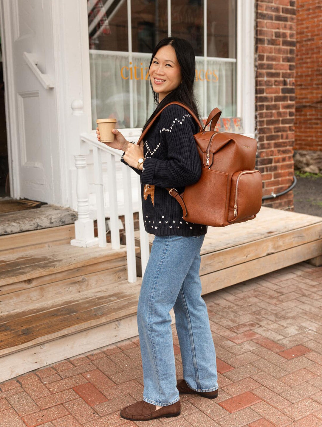 Poppy & Peonies The Cabin Backpack in Chestnut worn as a backpack by a woman holding a coffee, highlighting its structured shape and padded shoulder straps.