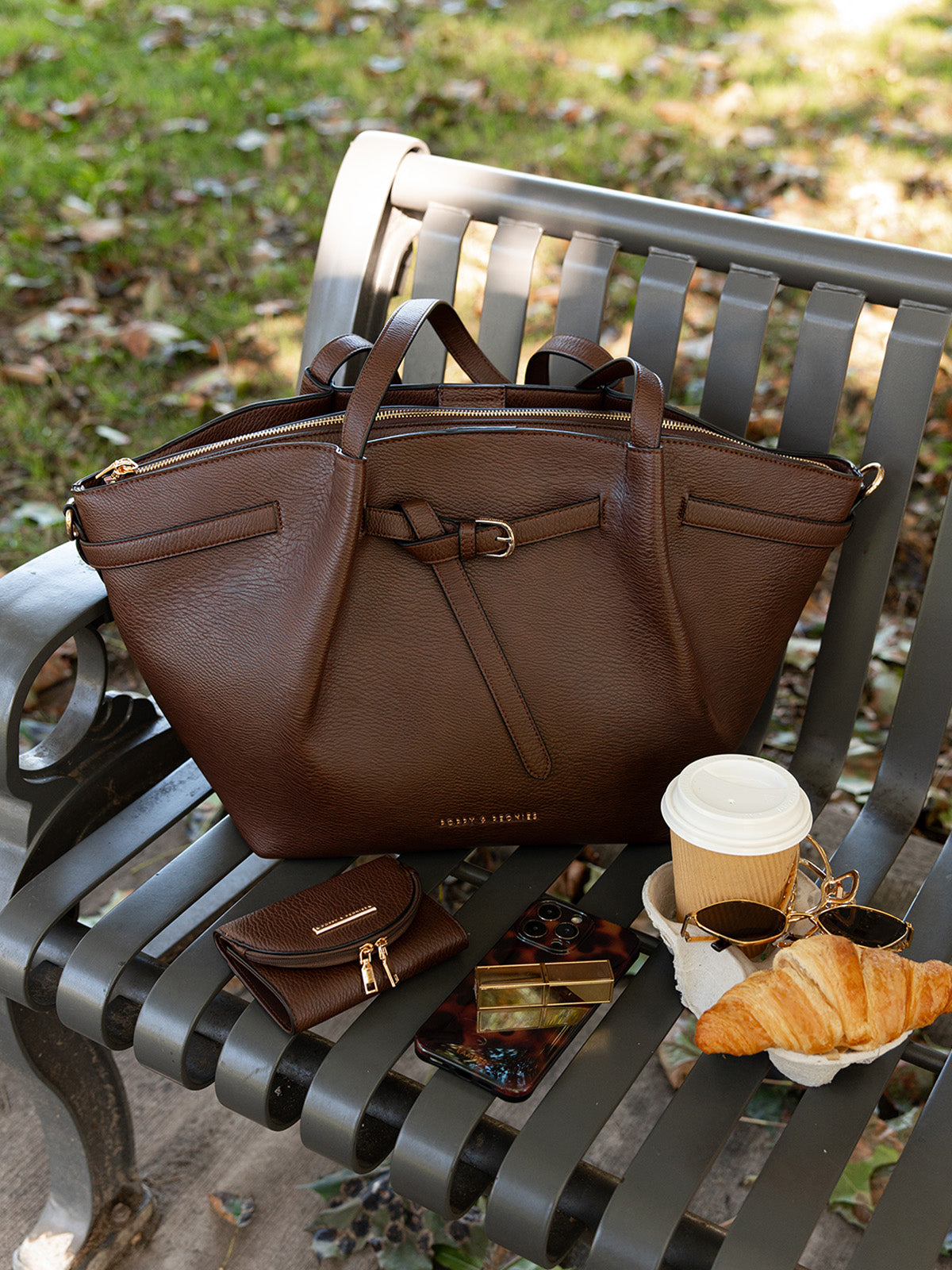 Poppy & Peonies Knot Tote in Espresso resting on a green park bench beside a matching accessory, takeaway coffee and autumn leaves.