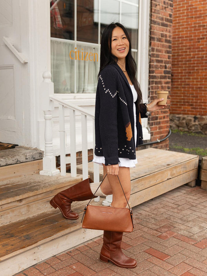 Poppy & Peonies The Soho Shoulder Bag in Chestnut carried by its slim strap by a woman standing on a brick sidewalk outside a café, wearing a navy cardigan and tall brown boots.