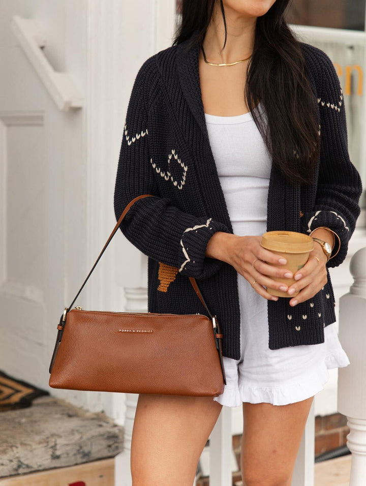 Poppy & Peonies The Soho Shoulder Bag in Chestnut worn over the shoulder with a navy knit cardigan and white shorts, styled with a takeaway coffee outside a storefront.