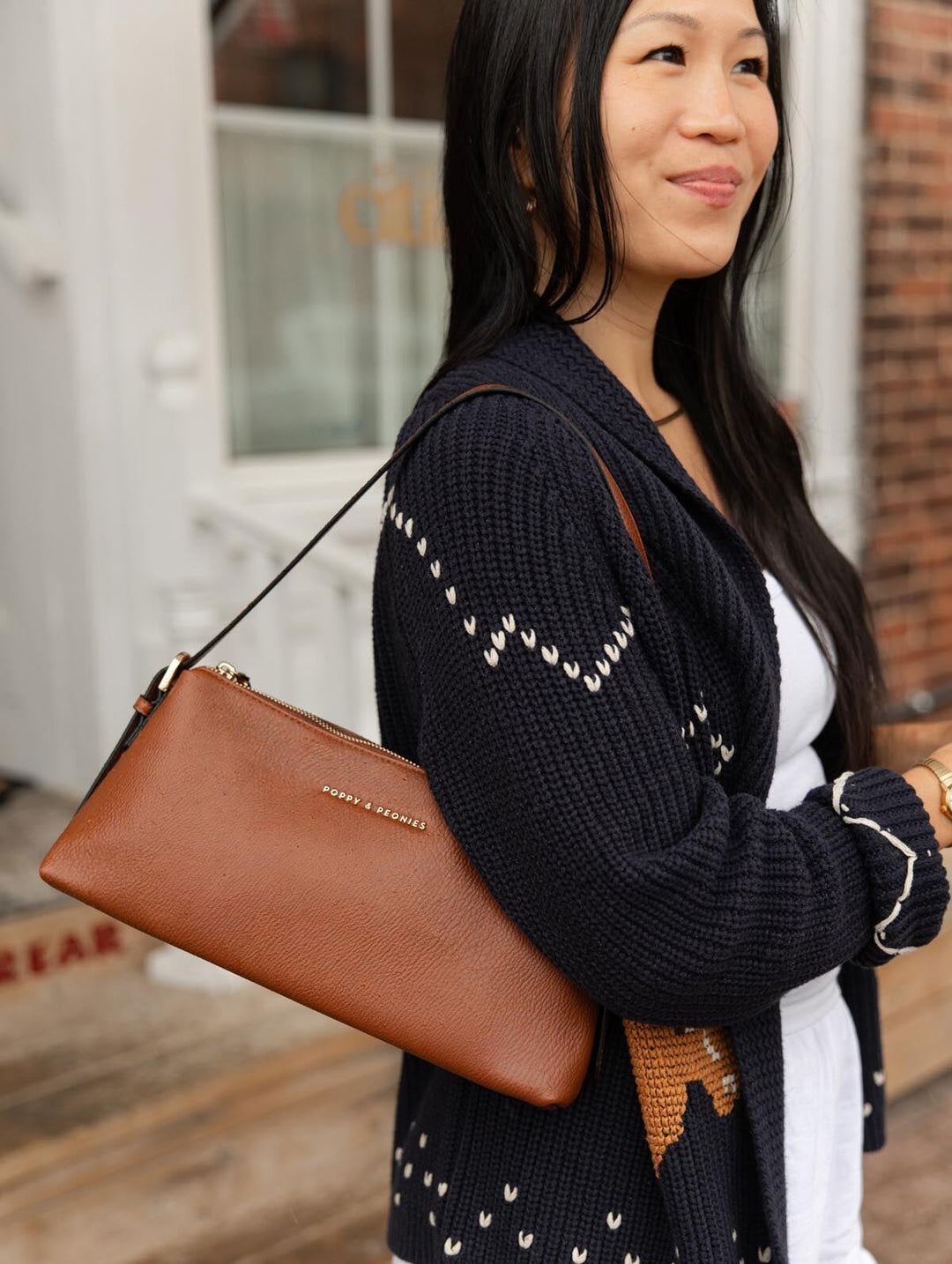 Poppy & Peonies The Soho Shoulder Bag in Chestnut worn over the shoulder by a woman in a navy cardigan and white outfit, showing the bag's slim profile.