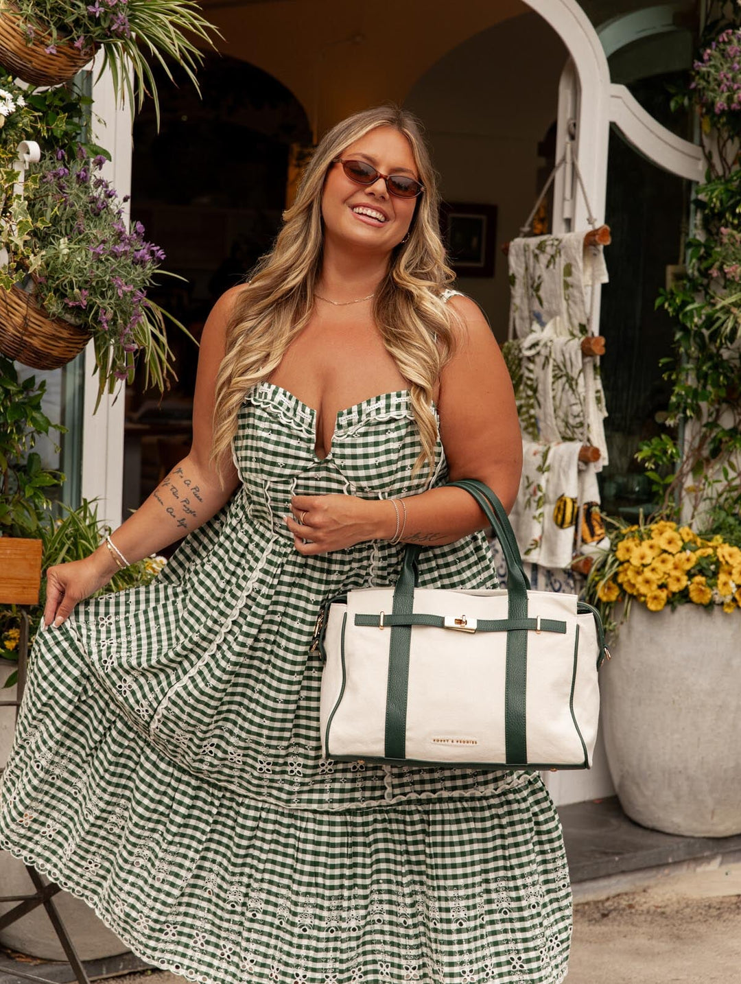 Woman in a green checkered dress holding a white and green handbag with floral decorations in the background.