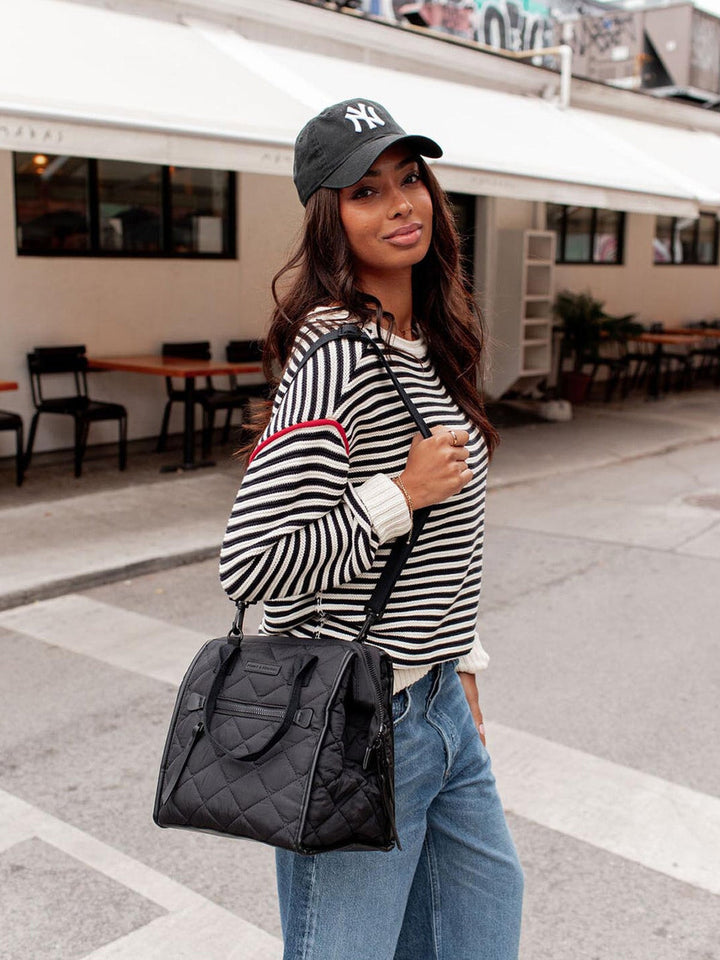 Woman wearing a striped sweater, cap, and jeans with a black lunch bag in an outdoor setting.