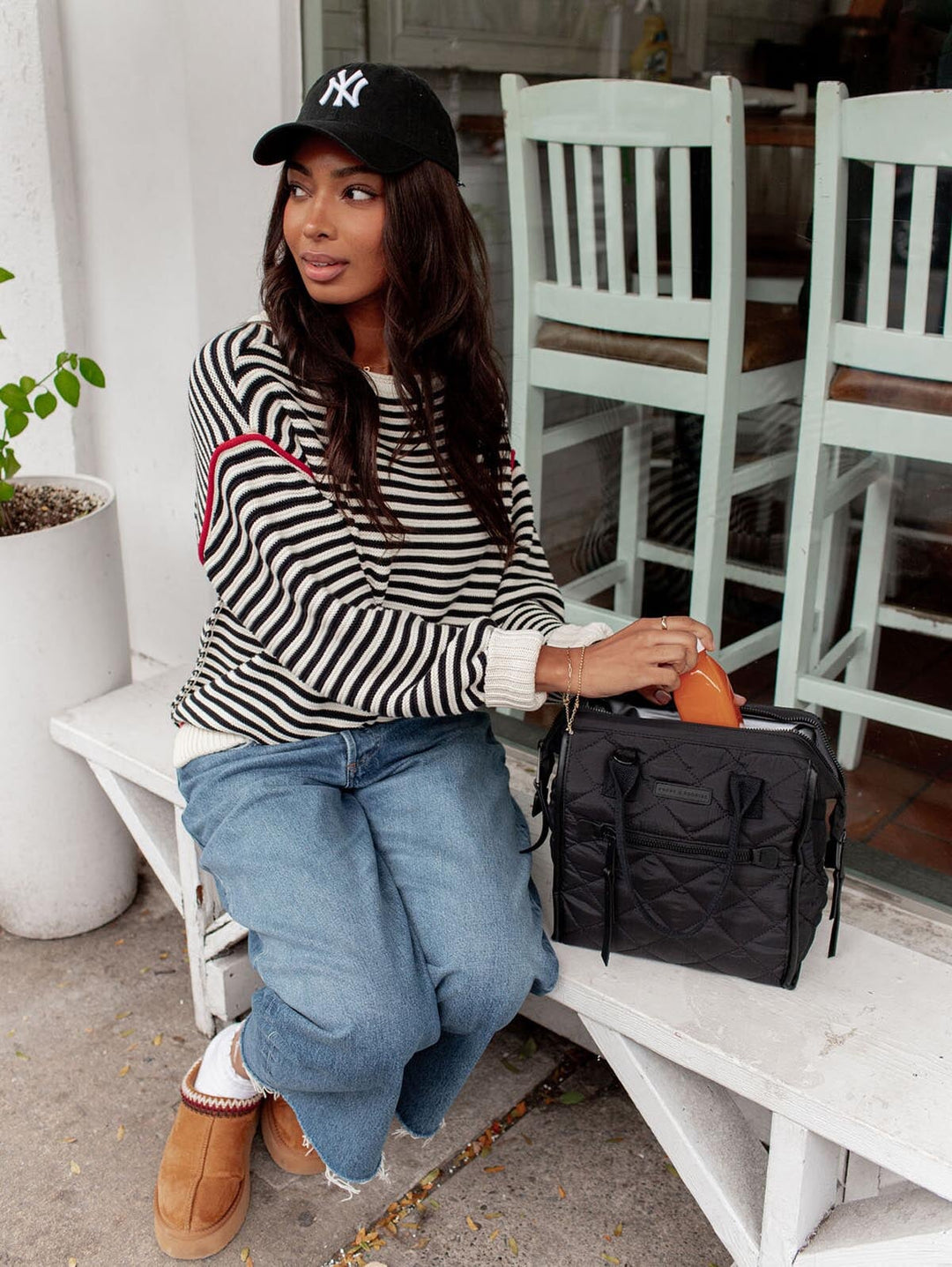 Woman sitting on a bench wearing a striped sweater and jeans, holding a black lunch bag.
