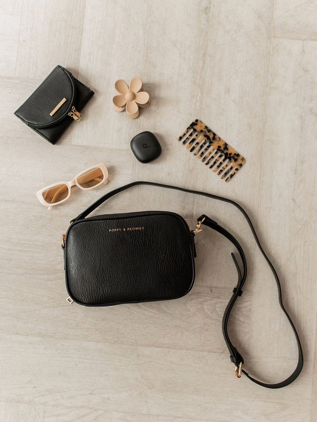 Black handbag with a strap, sunglasses, a small black bag, a flower, and a comb on a light wooden surface.