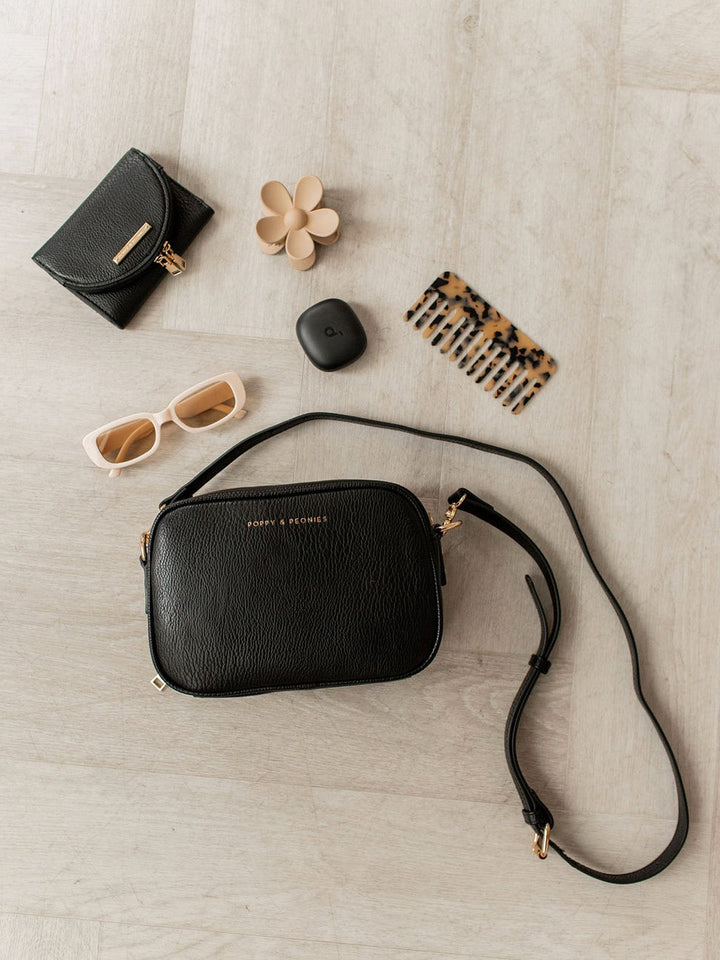 Black handbag with a strap, sunglasses, a small black bag, a flower, and a comb on a light wooden surface.