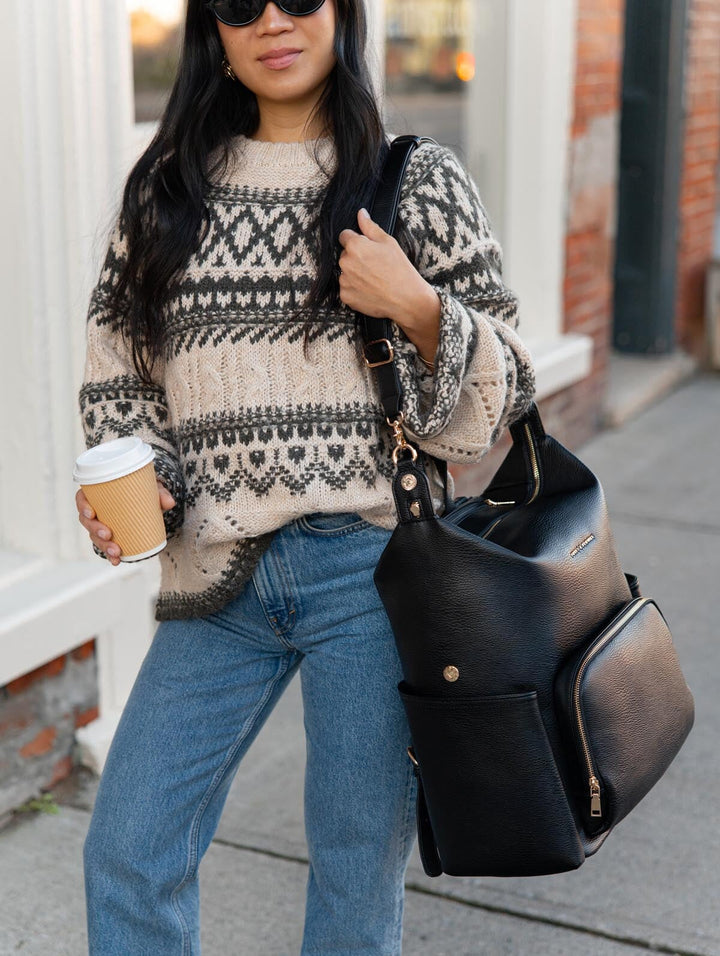 Poppy & Peonies The Cabin Backpack in Black carried over one shoulder by a woman holding a coffee, showing its spacious shape, top handle, and front organizer pocket.