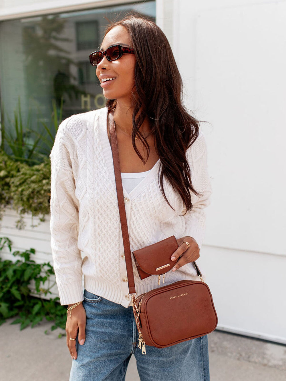 Woman wearing a white sweater, blue jeans, and sunglasses, holding a brown vegan leather crossbody bag.