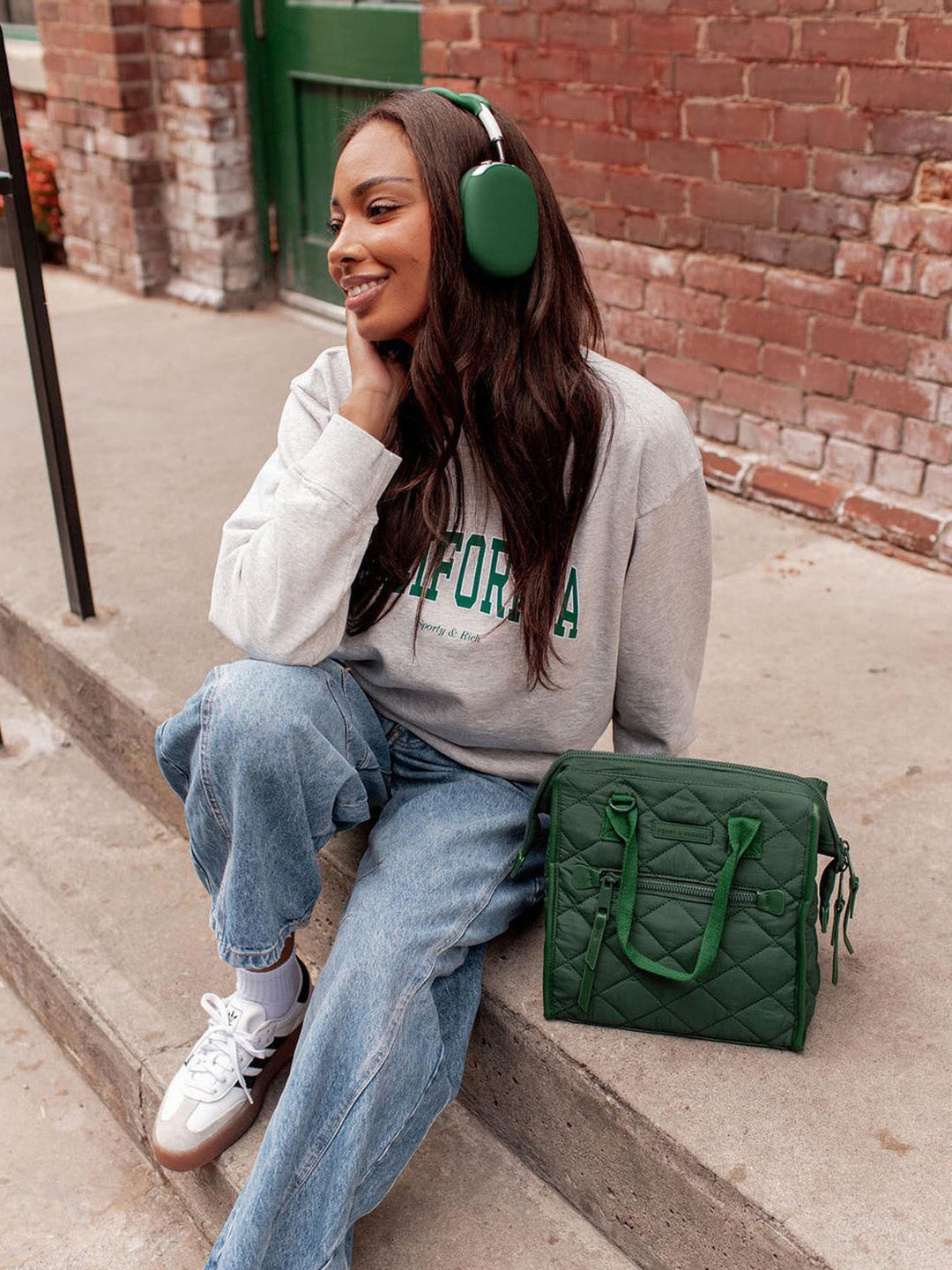 Woman sitting on steps wearing a grey sweatshirt with text, blue jeans, and green headphones, with a green lunch bag beside her.