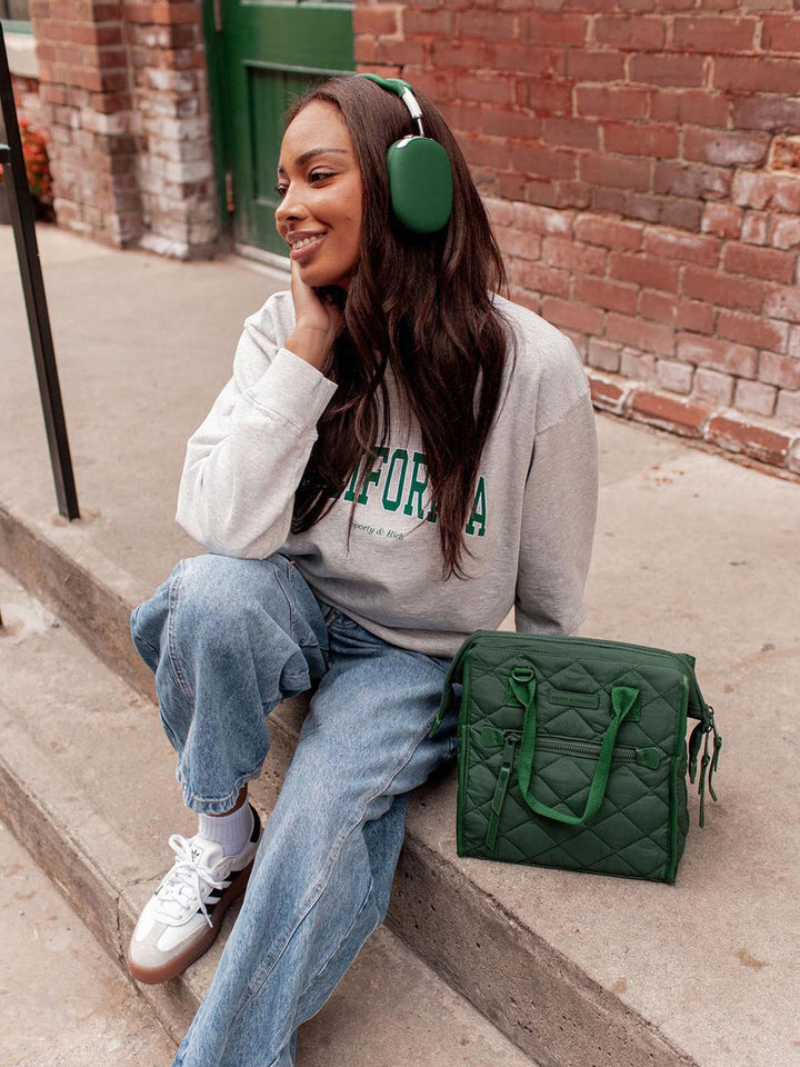 Woman sitting on steps wearing a grey sweatshirt with text, blue jeans, and green headphones, with a green lunch bag beside her.
