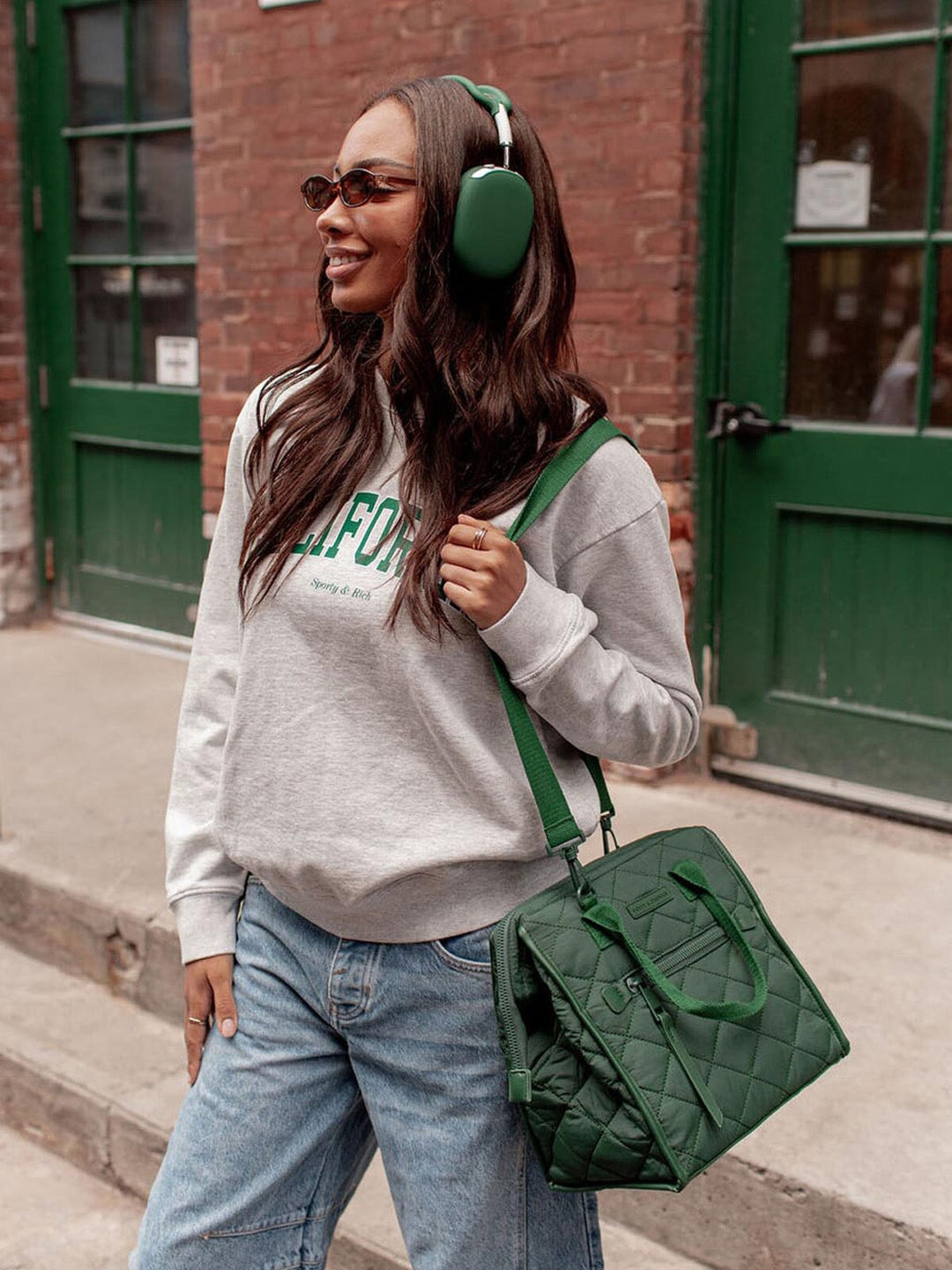 Woman wearing a grey sweatshirt and green headphones, holding a green lunch bag in front of a brick building.