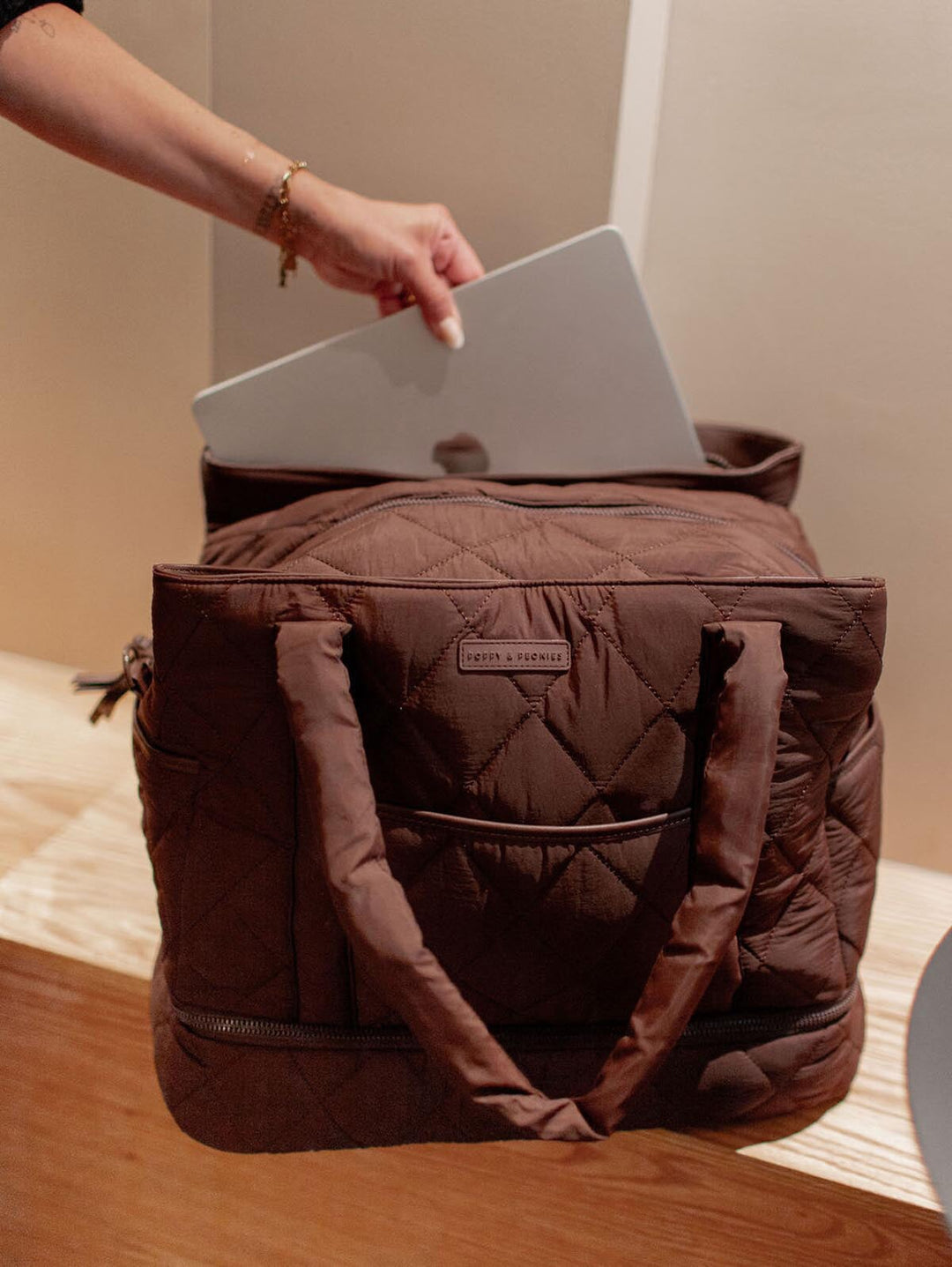Brown quilted bag with a person placing a laptop inside on a wooden surface.