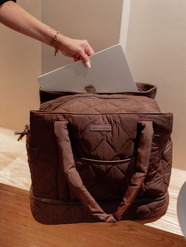 Brown quilted bag with a person placing a laptop inside on a wooden surface.