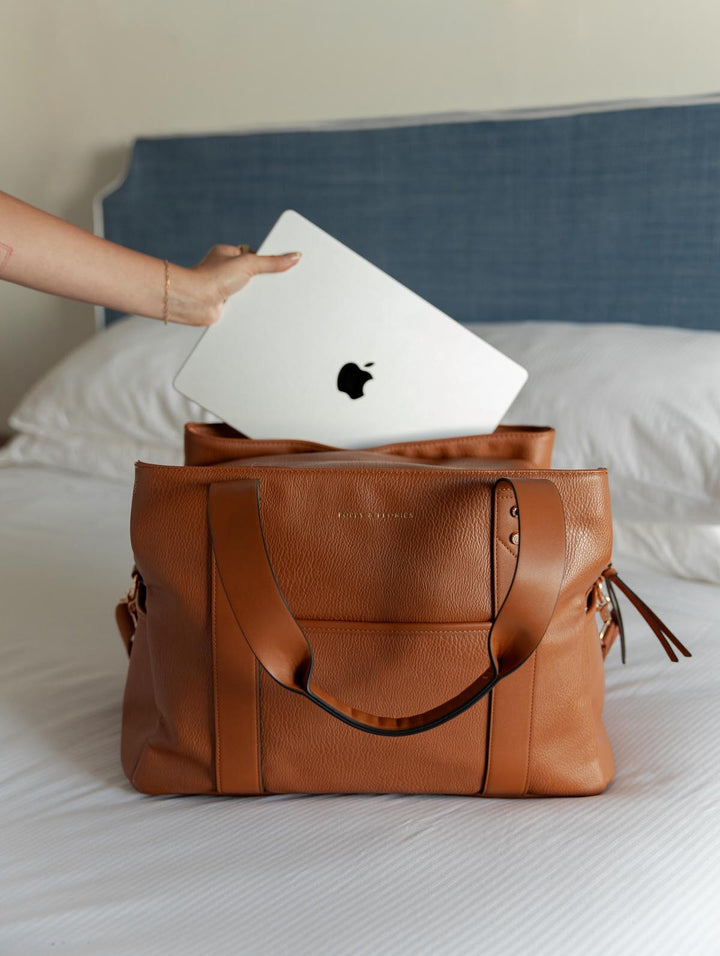 Brown vegan leather travel bag on a bed with an Apple laptop being placed inside