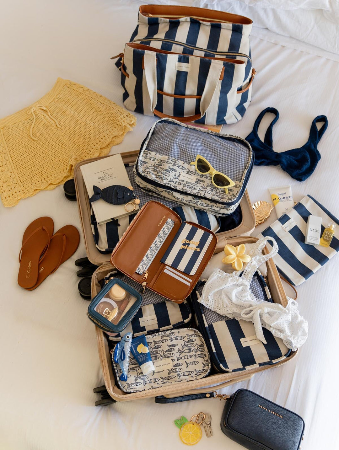 Open suitcase with travel items including packing cubes, a striped bag, sunglasses, and clothes on a bed.