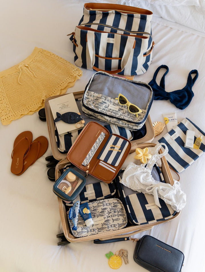 Open suitcase with travel items including packing cubes, a striped bag, sunglasses, and clothes on a bed.