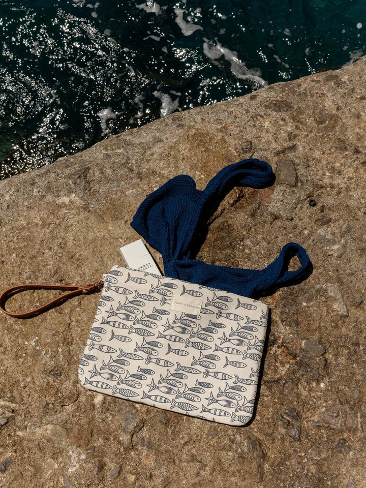 Small wet bag with a sardine pattern and blue drawstring on a stone surface near water.
