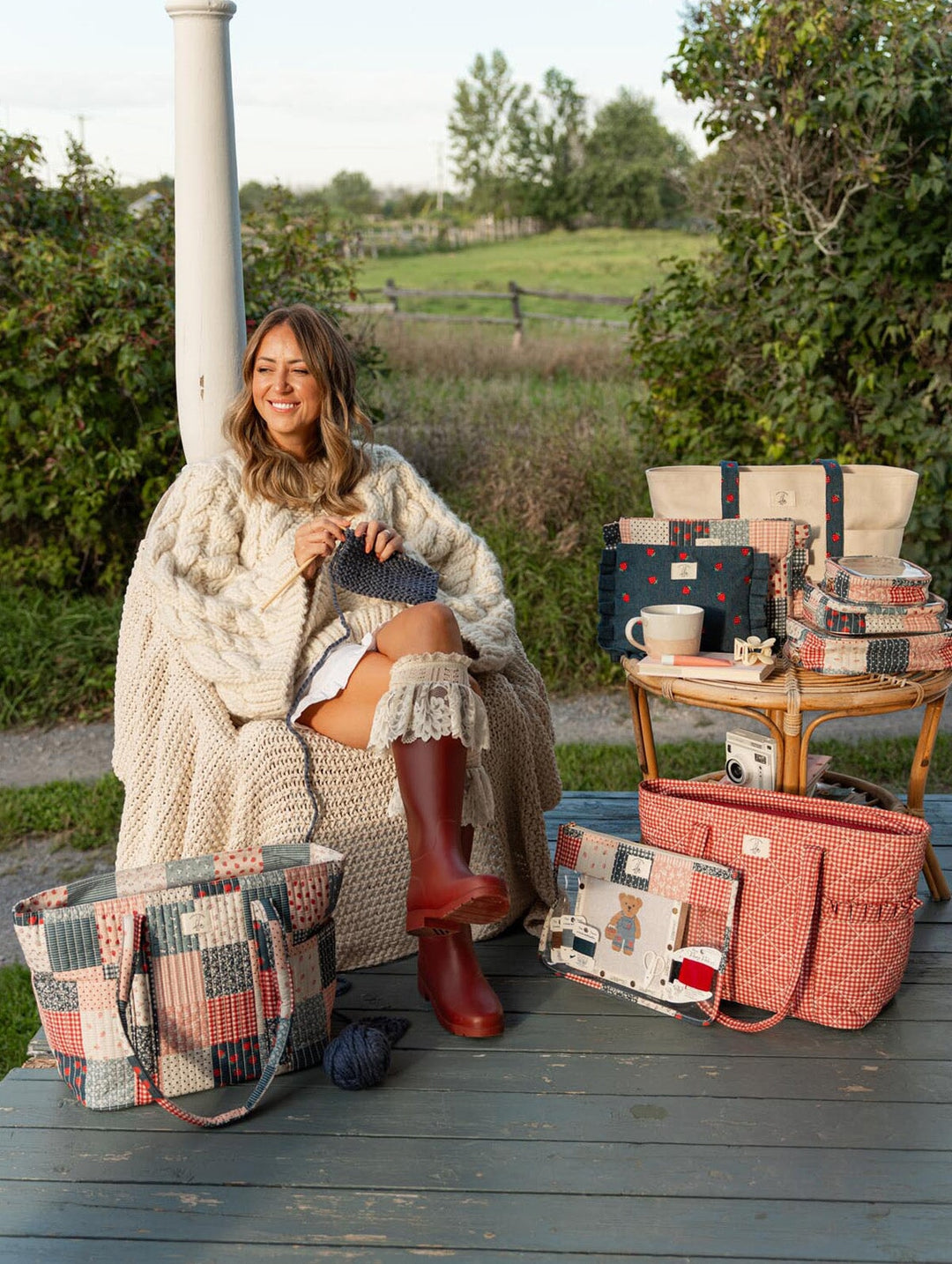 Poppy & Peonies Fallon Collection lifestyle image featuring Fallon knitting outdoors, surrounded by the Analog Tote in Strawberry Patchwork and Heart Berry Gingham, Strawberry Denim tote, coordinating organizers and craft accessories.