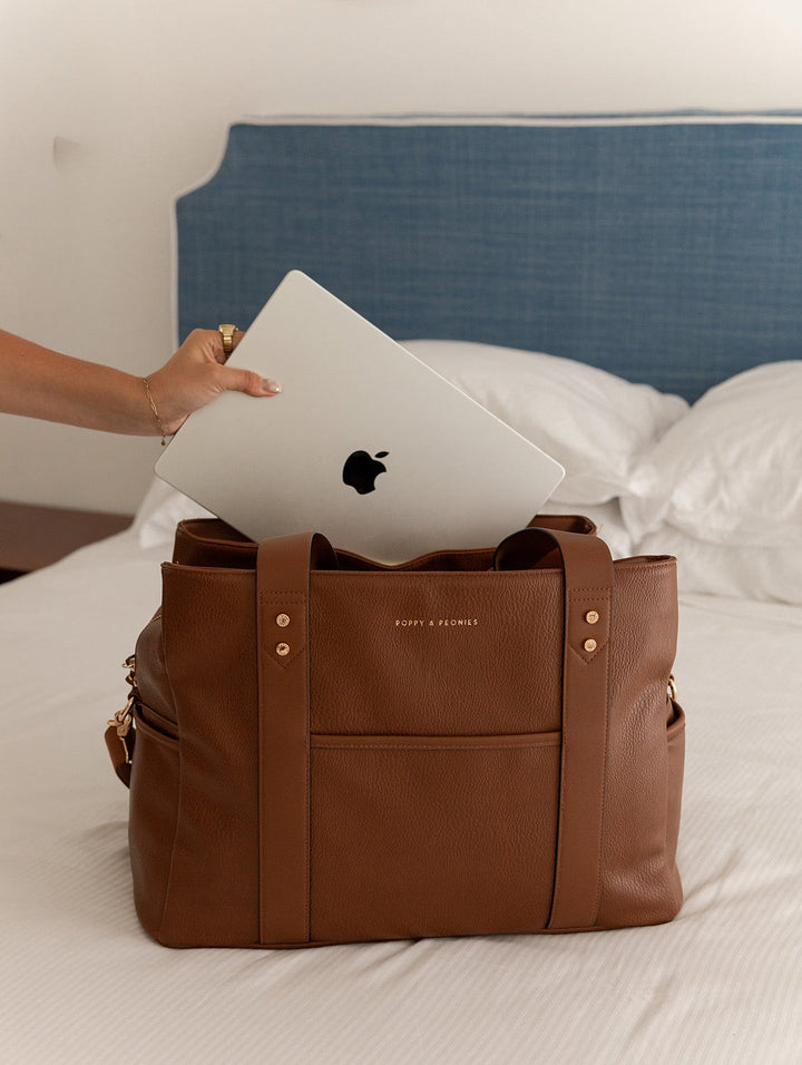 Brown vegan leather bag with a laptop being placed into it on a bed.