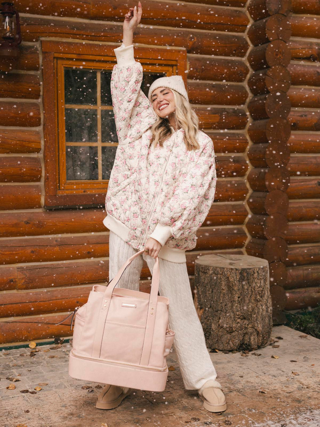 Woman in a floral sweater and white pants standing in front of a wooden cabin, holding a blush coloured vegan leather travel bag.
