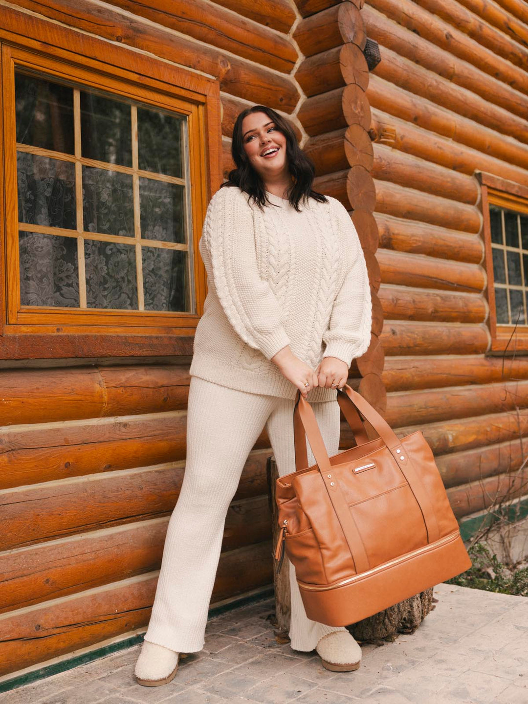 Woman holding a brown vegan leather bag in front of a wooden cabin