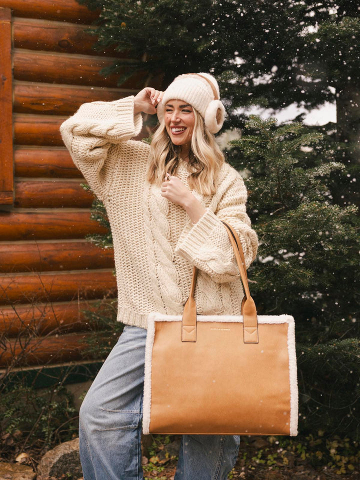 Woman in a beige knit sweater and hat holding a brown vegan leather bag outdoors.