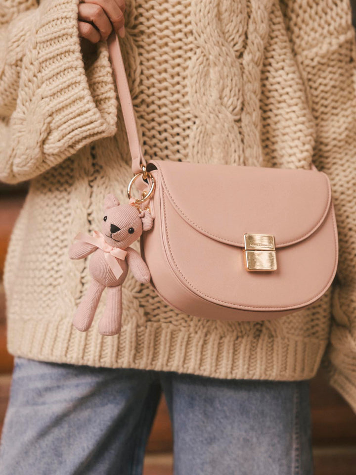 Pink crossbody bag with a teddy bear bag charm  held by a person wearing a beige knitted sweater.