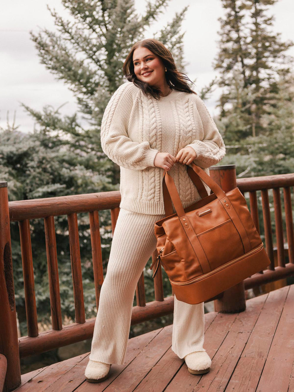 Woman in a cream sweater and pants holding a brown vegan leather travel bag on a wooden deck with trees in the background