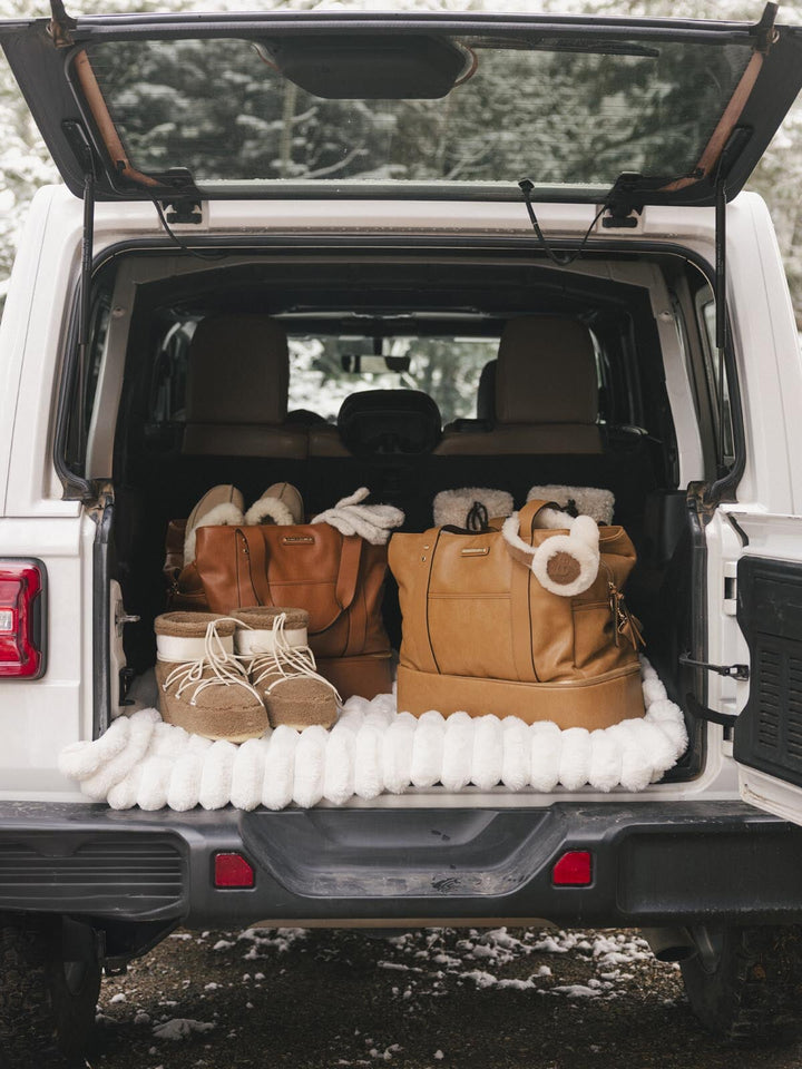 Trunk of a vehicle with vegan leather travel bags, winter boots and accessories on a snowy background.