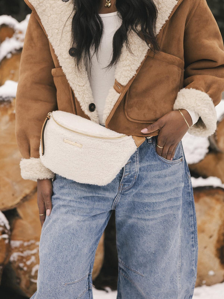 Person holding a cream faux fur belt bag, standing in a snowy landscape.