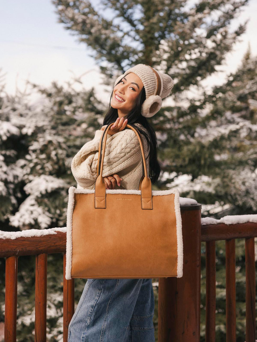 Person holding a brown vegan leather tote bag with a snow-covered pine tree background