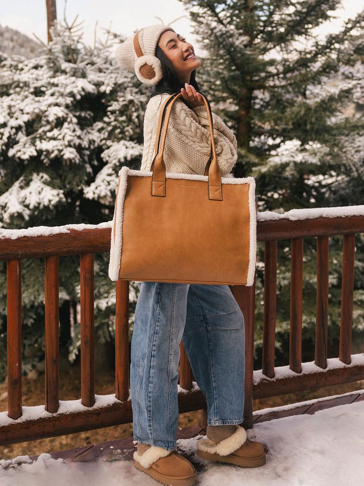 Person holding a brown vegan leather tote bag with white faux fur trim on a snowy deck.