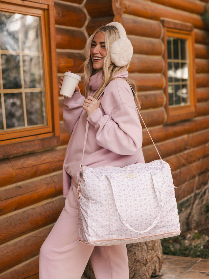 Woman in pink outfit with floral print duffel bag and ear muffs standing in front of a log cabin.