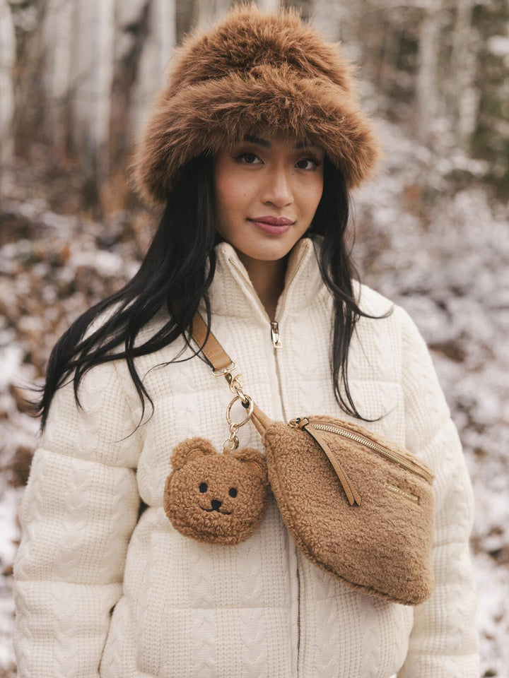 Woman in a snowy forest wearing a fur hat and white coat with a brown faux fur bag featuring a teddy bear charm.