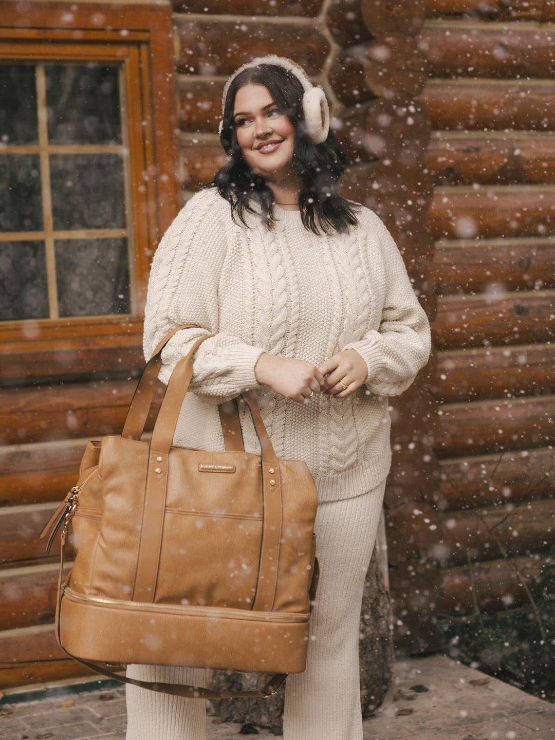 Woman in a cable knit sweater and white pants holding a brown vegan leather travel bag in front of a wooden cabin.