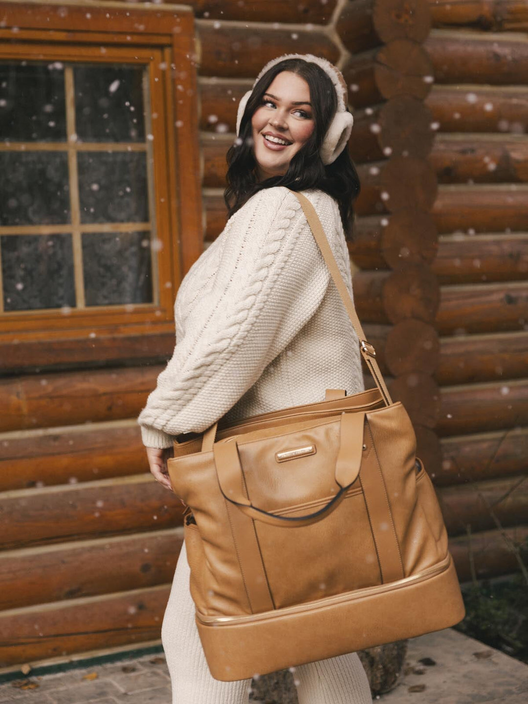 Woman holding a vegan leather travel bag in front of a log cabin