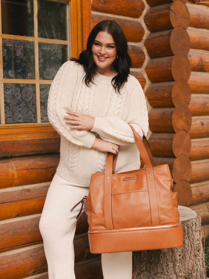 Woman in a white outfit holding a brown vegan leather bag in front of a wooden cabin.