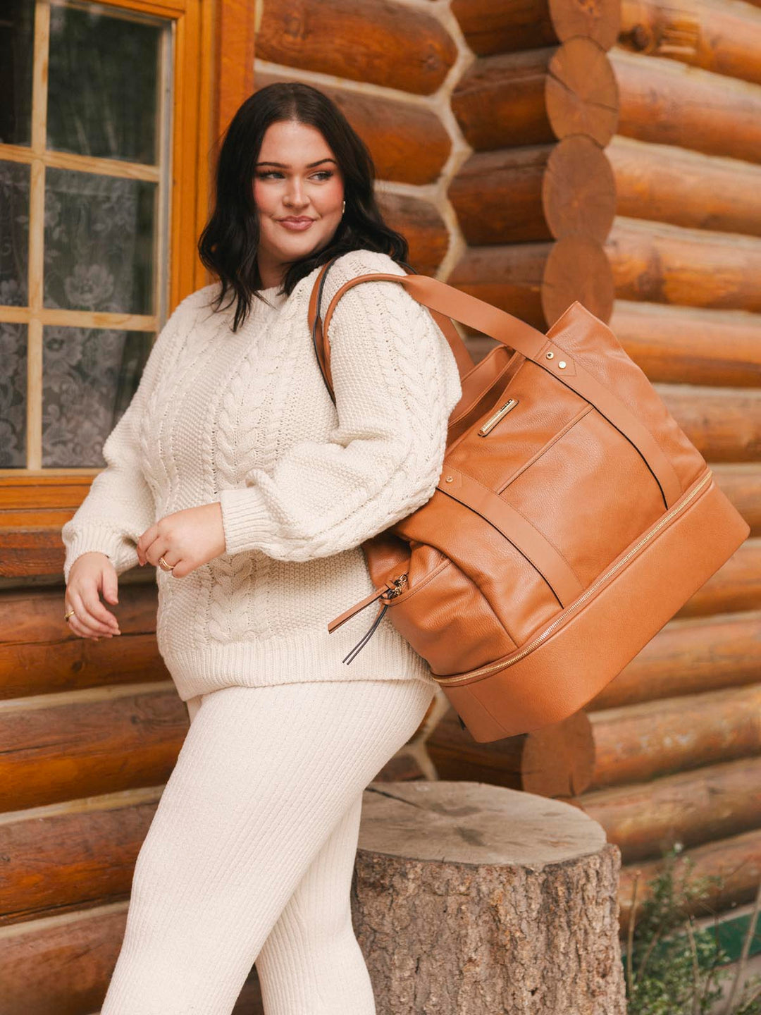 Woman in a white sweater with a brown vegan leather travel bag standing in front of a log cabin.