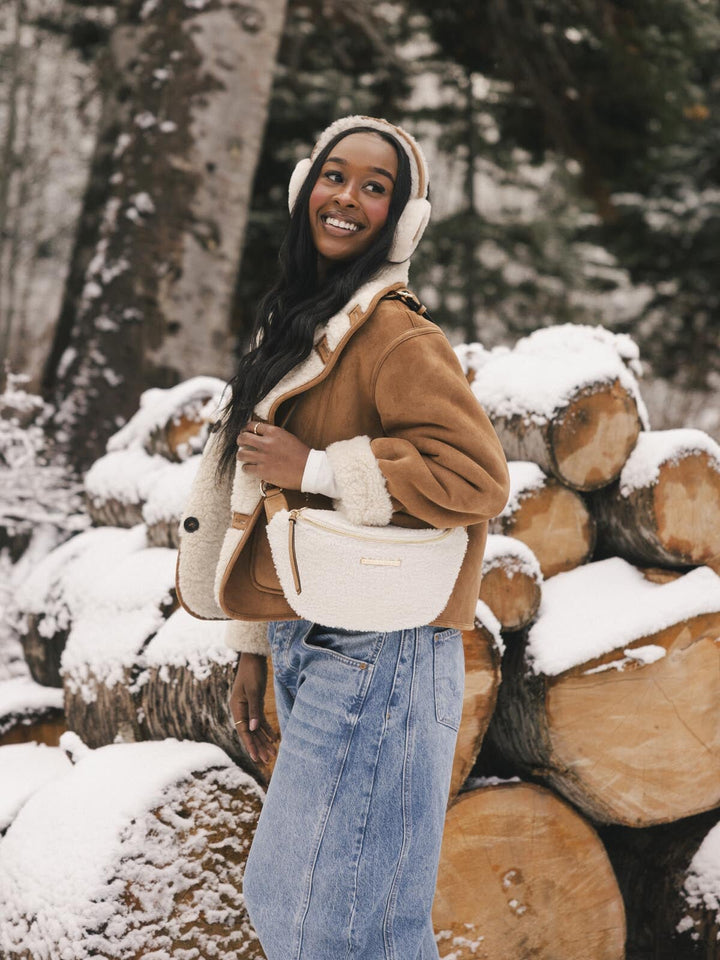 Woman with a cream faux fur crossbody bag standing next to a stack of logs in a snowy forest