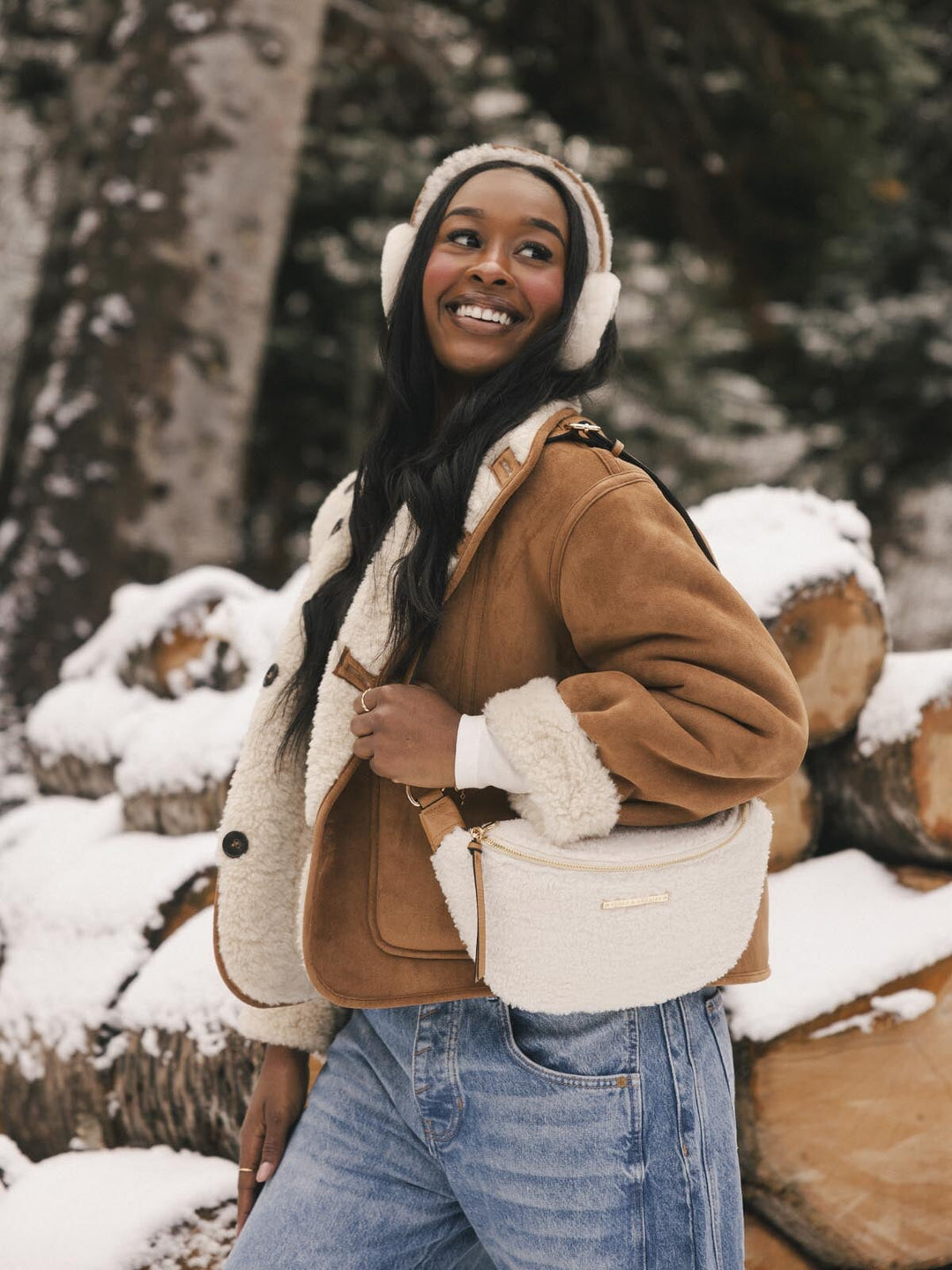 Person wearing a brown jacket with white fur lining, blue jeans, and carrying a cream faux fur bag in a snowy forest setting.