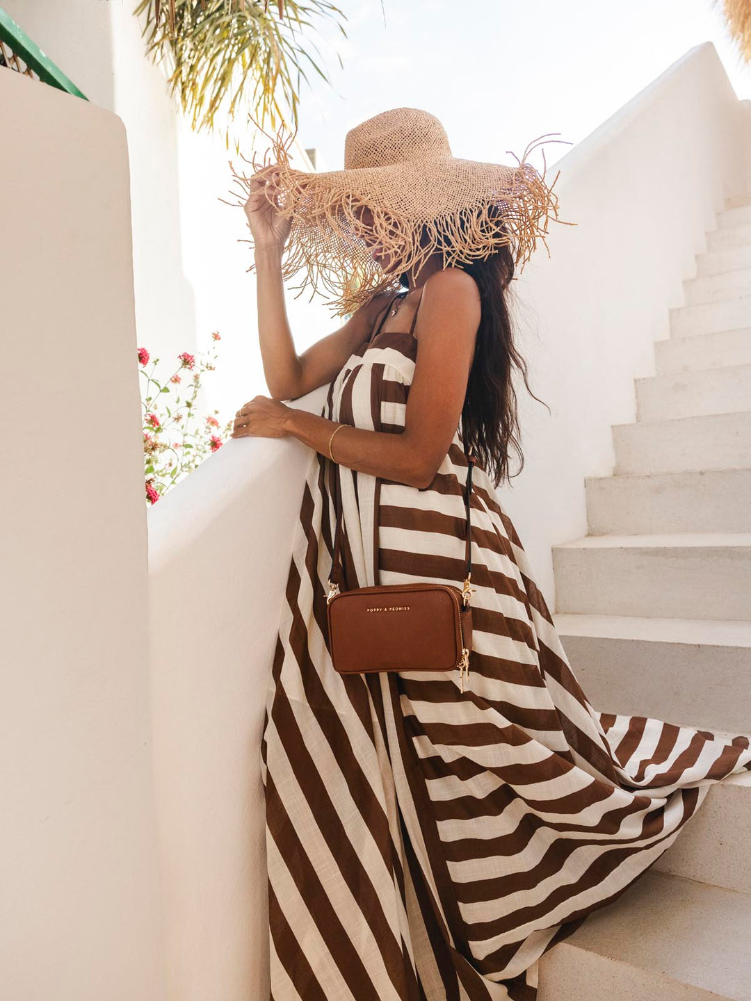 Woman in a striped dress and straw hat wearing a brown crossbody bag standing on a staircase.