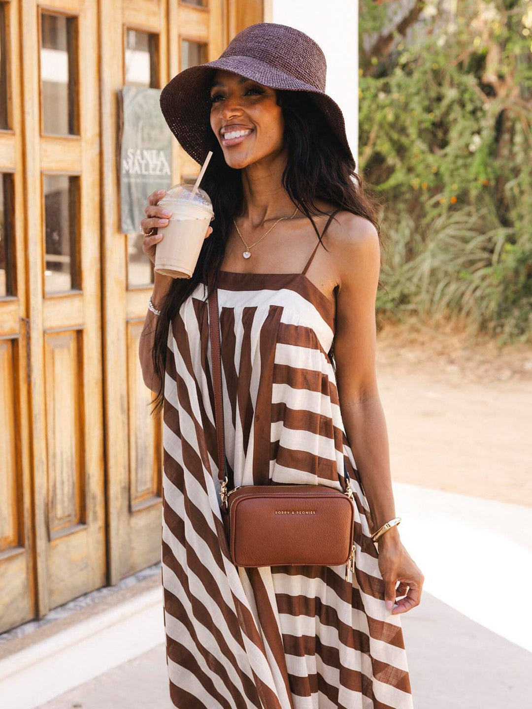 Woman in a brown and white striped dress and brown crossbody bag holding a drink, standing outdoors.
