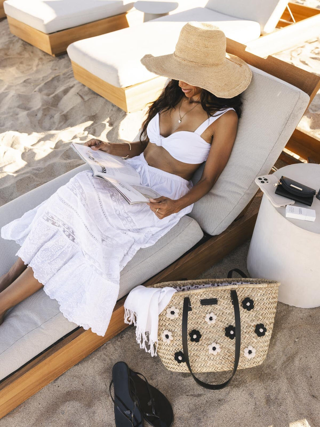 Woman in white dress and straw hat reading a magazine on a lounge chair with a handbag beside her.