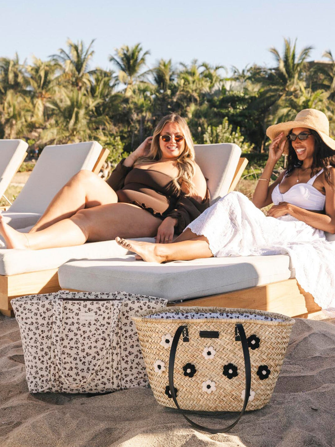 Two women lounging on beach chairs with a straw bag and striped tote bag in the foreground.