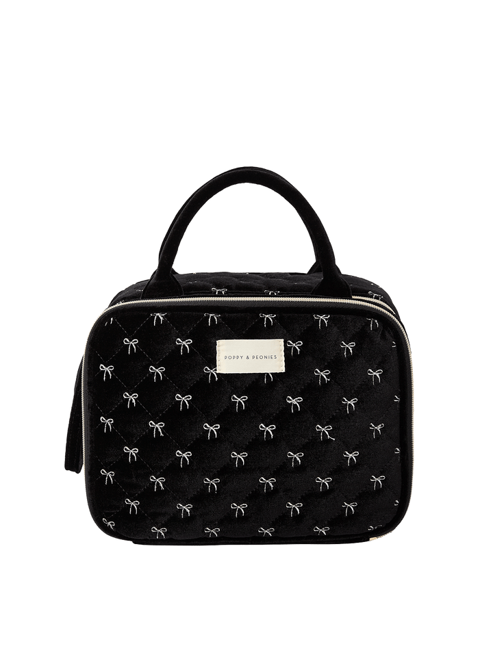 Poppy and Peonies The Routine Cosmetic Black Velvet featuring a structured rounded rectangular silhouette, soft black velvet exterior with delicate white bow patterns, dual black top handles, zip-around closure, and rectangular gold Poppy and Peonies logo plate on the front.