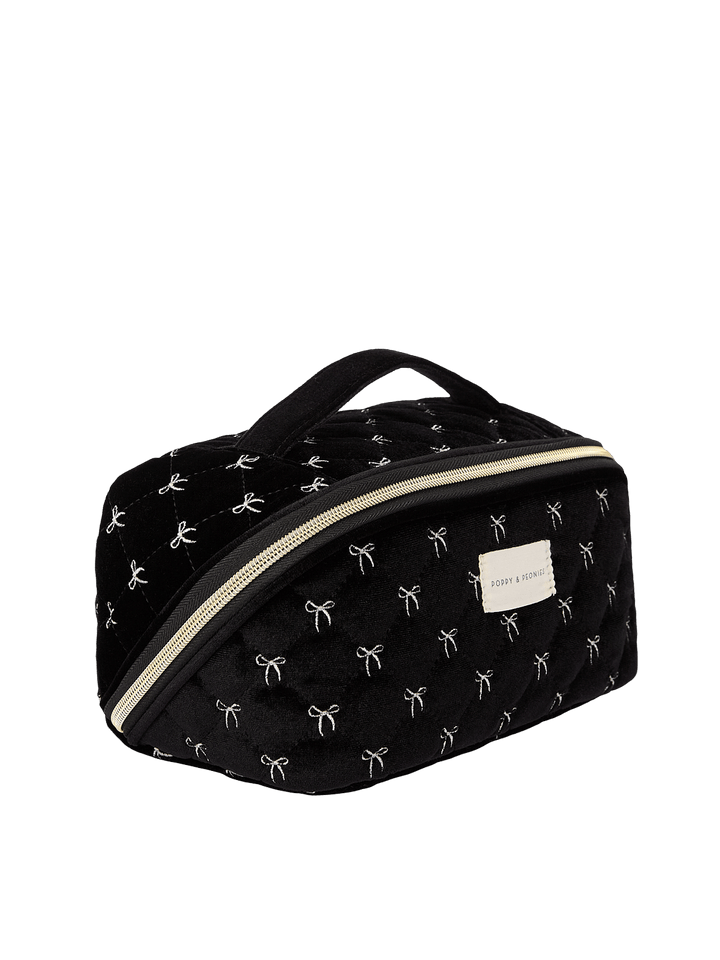 Poppy and Peonies Open and Glow Cosmetic Case Black Velvet displayed at a three-quarter angle, highlighting its structured rectangular shape, soft black velvet exterior with white bow patterns, top carrying handle, front zippered pocket, gold-tone hardware, and rectangular gold logo plate.