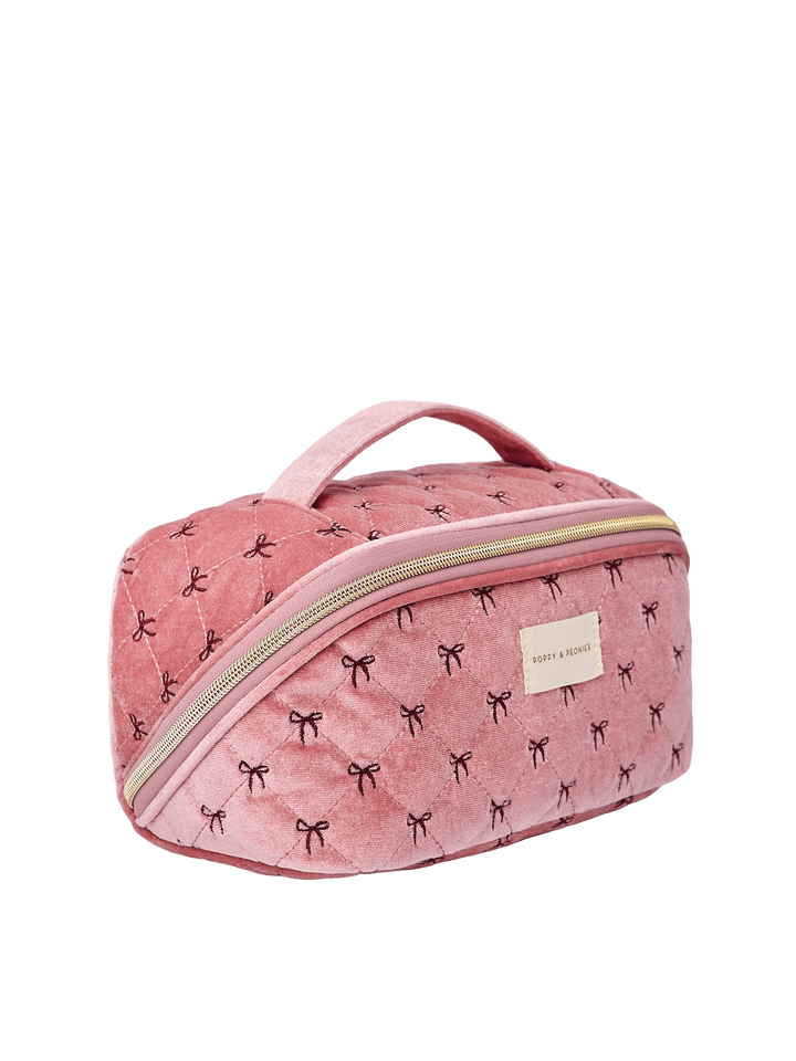 Poppy and Peonies Open and Glow Cosmetic Case Pink Velvet displayed at a three-quarter angle, highlighting its structured rectangular shape, soft pink velvet exterior with black bow patterns, top carrying handle, front zippered pocket, gold-tone hardware, and rectangular gold logo plate.