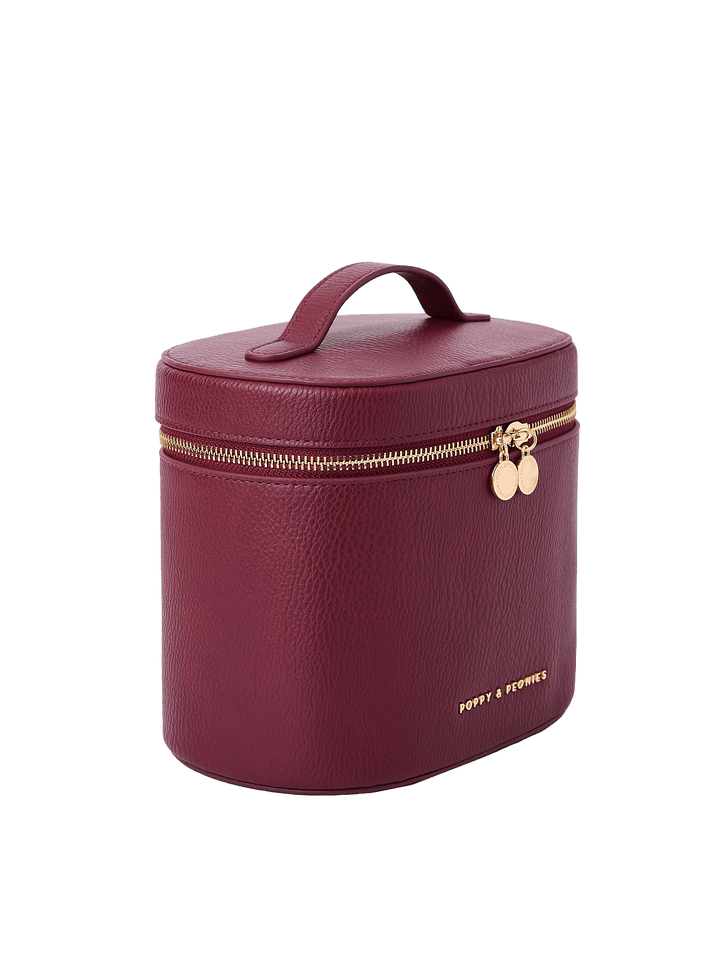 Poppy and Peonies The Vanity Case Berry displayed at a three-quarter angle, highlighting its structured silhouette, rich berry textured exterior, top carrying handle, gold-tone zipper hardware, and subtle Poppy and Peonies logo near the bottom.
