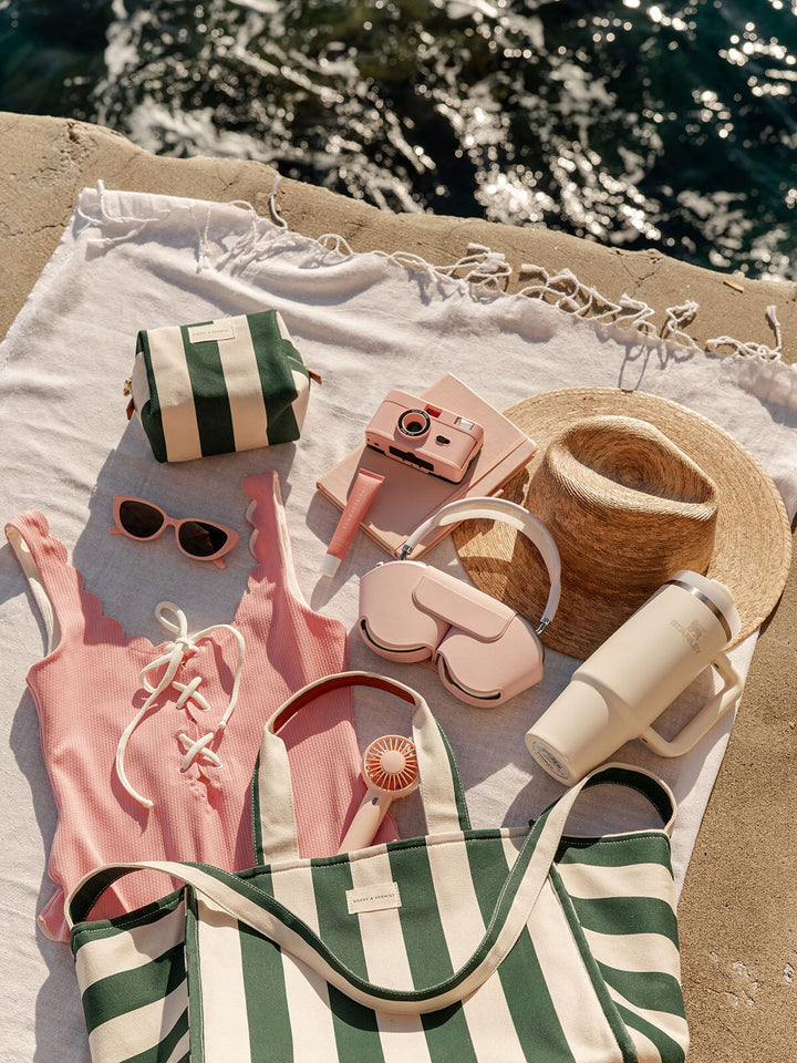 Beach accessories including a pink swimsuit, sunglasses, camera, green and white striped tote bag and hat on a towel by the water.