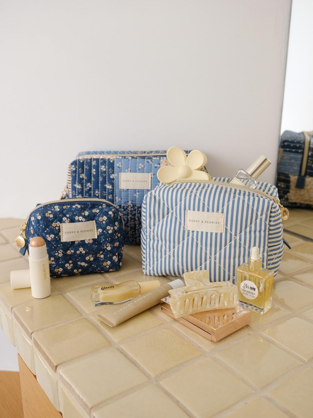 Product-focused lifestyle shot of the Cosmetic Trio in Navy Patchwork, featuring all three coordinating cosmetic cases in blue floral, striped, and patchwork prints styled alongside beauty essentials.